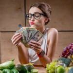 Une femme avec des lunettes compte des billets de banque, entourée de fruits et légumes frais sur une table.